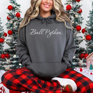 May include: A dark gray hooded sweatshirt featuring the white cursive text Ball Python. on the chest. The garment includes a drawstring hood and a front kangaroo pocket, worn with red and black buffalo plaid patterned lounge pants.