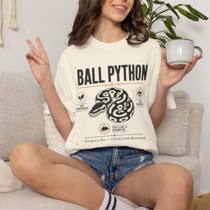 May include: An off-white short-sleeve t-shirt featuring a black graphic of a coiled ball python. The text reads BALL PYTHON, LOW MAINTENANCE, HIGH PERSONALITY, EATS LIKE A CHAMPION, and Not Just a Pet — A Scaly Little Roommate with small icons.
