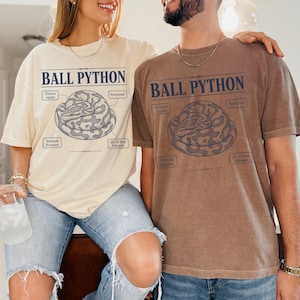 以下が含まれることがあります： クリーム色のTシャツと茶色のTシャツ。どちらも丸まったボールパイソンのグラフィックが描かれています。テキストには「BALL PYTHON」「Python regius」「Nocturnal」「Ambush Predator」「20-30 Year Lifespan」と記されています。両方のシャツのデザインは同一です。