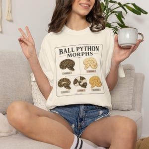 以下が含まれることがあります： BALL PYTHON MORPHSというタイトルのグラフィックプリントが施されたオフホワイトのTシャツ。Normal、Albino、Pastel、Pied、Bananaとラベル付けされた5匹のヘビのイラストが、細い黒い線で区切られたグリッド状に配置されています。