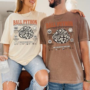 Könnte beinhalten: Ein cremefarbenes T-Shirt und ein braunes T-Shirt, beide mit einer Grafik eines zusammengerollten Königspythons und dem Text BALL PYTHON THE PERFECT COIL OF CHILL sowie Symbolen und Beschriftungen für Decken-Dieb, Nickerchen-Experte und pflegeleicht.