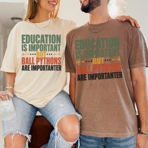 May include: A cream-colored short-sleeve t-shirt and a brown short-sleeve t-shirt, both featuring the text "EDUCATION IS IMPORTANT BUT BALL PYTHONS ARE IMPORTANTER" in green, yellow, and red block lettering with decorative arrow and star accents.
