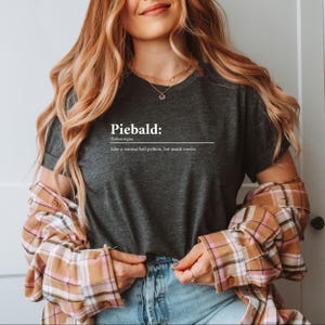 May include: Dark gray t-shirt with the word "Piebald" in white, followed by a definition and the phrase "Like a normal ball python, but much cooler." The model is wearing a plaid shirt over the t-shirt.