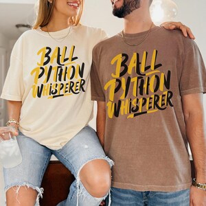 以下が含まれることがあります： クリーム色と茶色の半袖Tシャツ2枚。どちらも「BALL PYTHON WHISPERER」という文字が、太字でスタイリッシュな黄色と黒のフォントでプリントされています。Tシャツはクルーネックで、ゆったりとしたフィット感のデザインです。