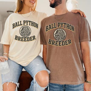 May include: Two short-sleeve crew neck t-shirts, one in cream and one in brown, each featuring the text BALL PYTHON BREEDER in a dark arched font above and below a circular graphic of a coiled snake, printed in black with a thin gold outline.