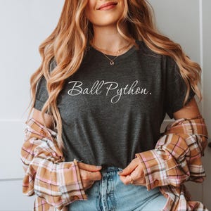 May include: A dark charcoal grey short-sleeved t-shirt featuring the white cursive text Ball Python. The shirt is worn under an unbuttoned tan, white, and pink plaid flannel shirt paired with light blue denim jeans, creating a layered casual look.