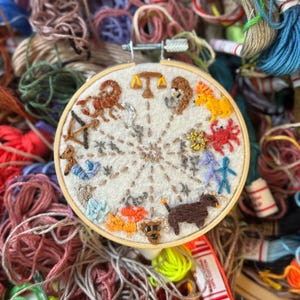 May include: A hand-stitched embroidery hoop featuring the twelve zodiac signs in colorful thread. The embroidery is set against a cream-colored fabric, surrounded by a variety of colorful embroidery floss.
