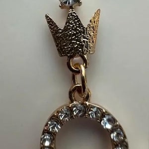 May include: Gold-toned earring with a crown design and a sparkling clear gemstone at the top. The earring features a small crown with a textured surface and a circular design adorned with multiple clear gemstones.