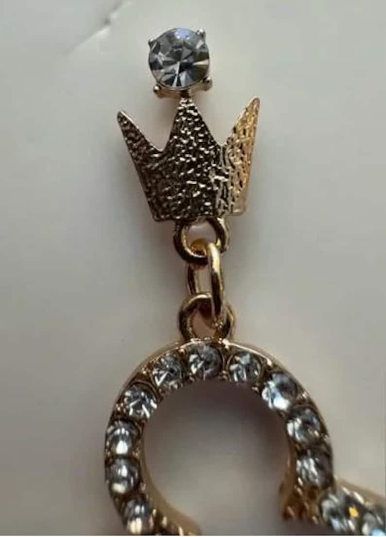 May include: Gold-toned earring with a crown design and a sparkling clear gemstone at the top. The earring features a small crown with a textured surface and a circular design adorned with multiple clear gemstones.