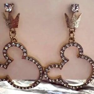 May include: A pair of gold-toned earrings featuring a Mickey Mouse silhouette design. The earrings are outlined with small, clear crystals and topped with a crown and a single clear crystal. The earrings are designed for pierced ears.