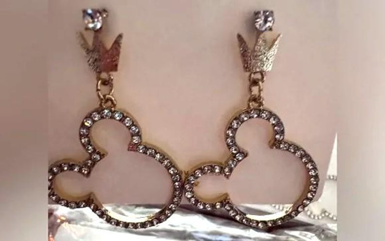 May include: A pair of gold-toned earrings featuring a Mickey Mouse silhouette design. The earrings are outlined with small, clear crystals and topped with a crown and a single clear crystal. The earrings are designed for pierced ears.