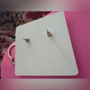 May include: A pair of gold-colored stud earrings with a post back, displayed on a white earring card. The earrings are small and round, with a simple design. The card is set against a pink background.