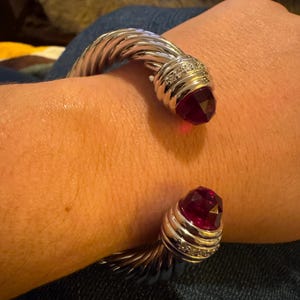 Peut inclure: Bracelet manchette argenté avec un design torsadé. Le bracelet présente à chaque extrémité une pierre précieuse rouge foncé facettée, sertie dans une lunette, ornée de petites pierres claires. Le bracelet est porté sur un poignet.