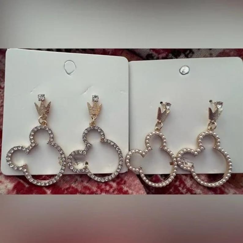May include: Two pairs of Mickey Mouse-shaped earrings. One pair is outlined with clear rhinestones, and the other is outlined with faux pearls. Both pairs have a gold-colored crown at the top.