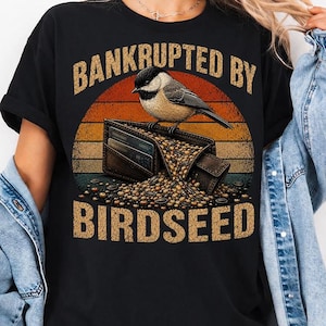 May include: A black t-shirt featuring a graphic of a chickadee perched on an open wallet overflowing with birdseed. The design includes a sunset-coloured circular background and the text "BANKRUPTED BY BIRDSEED" in a bold, vintage-style font.