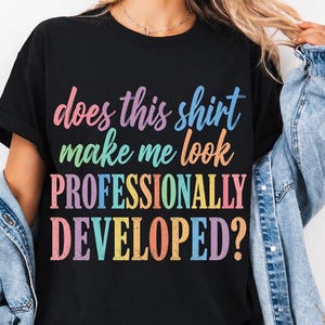 Peut inclure: Un t-shirt noir à manches courtes avec le texte multicolore does this shirt make me look PROFESSIONALLY DEVELOPED? Le lettrage utilise une variété de couleurs pastel et vives, notamment le rose, le bleu, le jaune et l'orange, dans une police décontractée.
