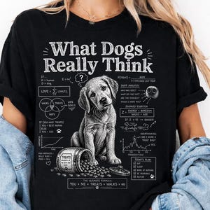 Könnte beinhalten: Schwarzes T-Shirt mit dem weißen Aufdruck "What Dogs Really Think". Auf der Vorderseite befindet sich eine Grafik eines Hundes mit Leckerlis und Diagrammen. Das Shirt besteht aus einem weichen Material.