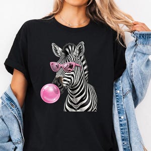 Op de afbeelding: Zwart t-shirt met een afbeelding van een zebra met een roze zonnebril en een roze bubbel. De zebra heeft zwart-witte strepen. Het t-shirt heeft korte mouwen.