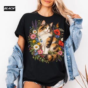 Puede incluir: Una camiseta negra con un estampado gráfico de un gato calicó sentado en medio de un colorido arreglo de varias flores y mariposas. La palabra BLACK está impresa en la esquina superior izquierda en fuente blanca sobre fondo negro.