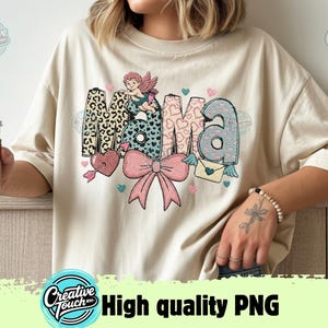 Peut inclure: T-shirt crème avec le mot "MAMA" dans un design décoratif. Le "M" a un imprimé léopard, le "A" est bleu avec des cœurs, et les autres lettres ont un thème de cœur et de nœud. Un petit cupidon est perché au-dessus.