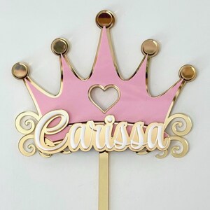 May include: A cake topper featuring a pink crown shape with a gold border and five gold spheres at the points. A heart cutout sits in the center. The name Carissa is written in white script with a gold outline across the front. A gold stake extends below.