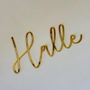 May include: A gold-colored metal sign that spells out the word "Hello" in a cursive font. The sign is made of a shiny, reflective material and is mounted on a white wall. The word is written in a continuous line.