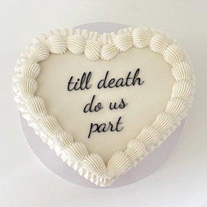 May include: A heart-shaped cake with white frosting and decorative piped edges. The cake features the phrase "till death do us part" in black script, making it suitable for weddings or anniversaries.