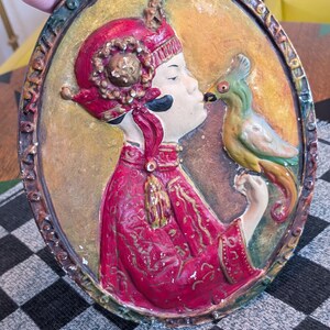 May include: Oval-shaped relief sculpture featuring a figure in a red and gold outfit kissing a green parrot. The sculpture has a gold background and a decorative border. The figure is wearing a red hat with gold accents.