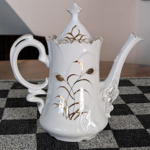 May include: White porcelain teapot with a gold wheat stalk design. The teapot has a curved handle, a spout, and a lid with a finial. The edges of the teapot and lid are trimmed with gold. A decorative piece for serving tea.