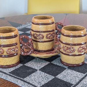 May include: Three ceramic mugs shaped like barrels with a textured tan finish and dark brown horizontal bands. Each mug features a handle moulded into the shape of a pretzel with white speckles. The mugs are arranged around a matching ceramic saucer.
