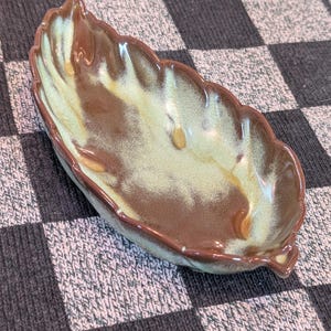 May include: A leaf-shaped ceramic dish with a scalloped edge. The dish features a blend of brown, tan, and green hues. The rim is a darker brown, and the interior has a mottled appearance. This decorative dish is suitable for serving snacks or as a decorative accent.