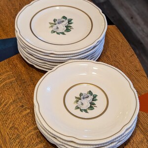 May include: A stack of white porcelain plates with a gold rim and a floral design. The plates have a scalloped edge and a gold border around the floral center. The floral design features white roses and green leaves.