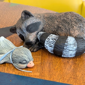 May include: A ceramic raccoon figurine with a sleeping pose, featuring a black and white striped tail and a gray and brown body. A smaller, sleeping bird figurine with a blue ribbon and pink beak is also present.
