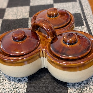 May include: A three-section ceramic condiment server with a handle. Each section has a rounded shape with a lid and a small knob. The server is brown with a cream-colored base. The handle is also brown.