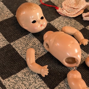 May include: A disassembled vintage doll with a peach-colored head, torso, and limbs. The doll's head has black eyes and a red mouth. The doll parts are arranged on a gray and black checkered surface.