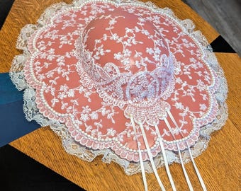 May include: A coral-colored sun hat with a delicate floral pattern and white lace trim. The hat features a wide brim and is adorned with white ribbons. The design is intricate and feminine, suitable for special occasions or vintage-inspired fashion.