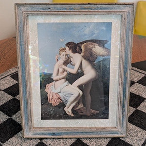 Puede incluir: Una lámina enmarcada que representa una escena clásica de una figura alada besando a una persona sentada. La obra presenta tonos suaves de azul y rosa, dentro de un marco de madera desgastada en azul claro y gris con un paspartú de color blanco roto.