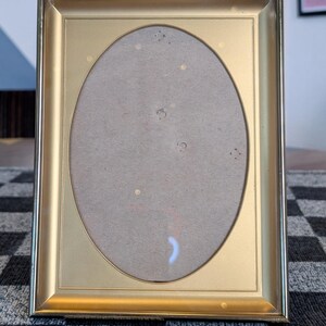 May include: A rectangular gold-colored picture frame with an oval opening. The frame has a simple, elegant design and is made of a shiny, metallic material. The frame is empty, with a gray backing visible inside.