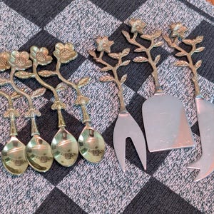 May include: A set of four gold-toned metal spoons and three serving utensils, including a cheese fork, a flat spreader, and a cheese knife. Each piece features a decorative handle shaped like a flowering branch with textured leaves and petals.