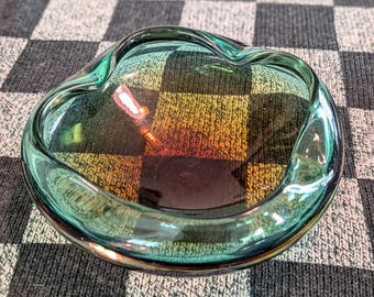 May include: A round, shallow glass bowl featuring a translucent teal rim that transitions into a warm amber and brown center. The glass has a smooth, organic, undulating edge and a polished finish, creating a reflective surface.