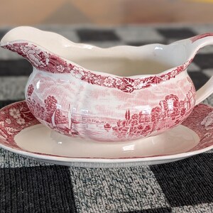 May include: A white and red gravy boat with a matching saucer. The boat and saucer feature a scenic landscape design with sailboats and buildings. The rim of the boat is decorated with a floral pattern.