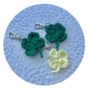 May include: Three crochet shamrock keychains. Two are green, and one is pale yellow. Each shamrock has a silver-colored clasp. The keychains are on a textured, light blue background.
