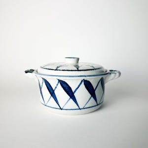 May include: A white ceramic lidded casserole dish featuring a hand-painted blue diamond pattern around the exterior. The round vessel includes two small side handles and a matching lid with a central knob, all finished with a subtle crackle glaze.