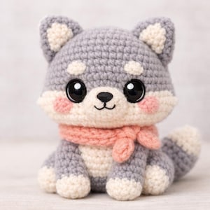 Puede incluir: Un juguete de perro Shiba Inu tejido a ganchillo. El perro es gris con detalles blancos y rosas, incluyendo una bufanda rosa. Tiene grandes ojos negros y mejillas rosas. El juguete es hecho a mano.
