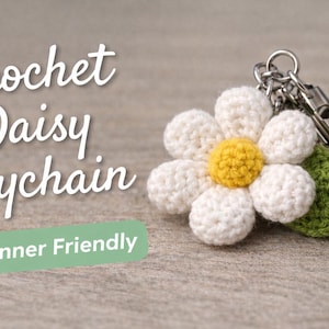 Op de afbeelding: Een gehaakte sleutelhanger in de vorm van een madeliefje met witte bloemblaadjes en een gele kern. De sleutelhanger heeft een groen blad en een zilverkleurige metalen ring. De tekst op de afbeelding luidt "Crochet Daisy Keychain" en "Beginner Friendly".