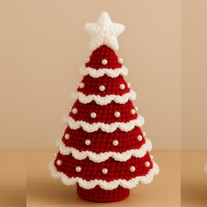Può includere: Decorazione per albero di Natale fatta a mano. L'albero è all'uncinetto con filo rosso, con bordi smerlati bianchi e piccole perle bianche. Una stella bianca in cima all'albero, creando una decorazione festiva.