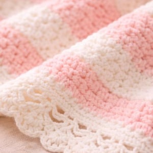May include: Close-up of a handmade crochet blanket with alternating stripes of soft pink and white. The edge features a delicate, scalloped trim. The blanket is made of a soft yarn, perfect for a baby or as a decorative throw.