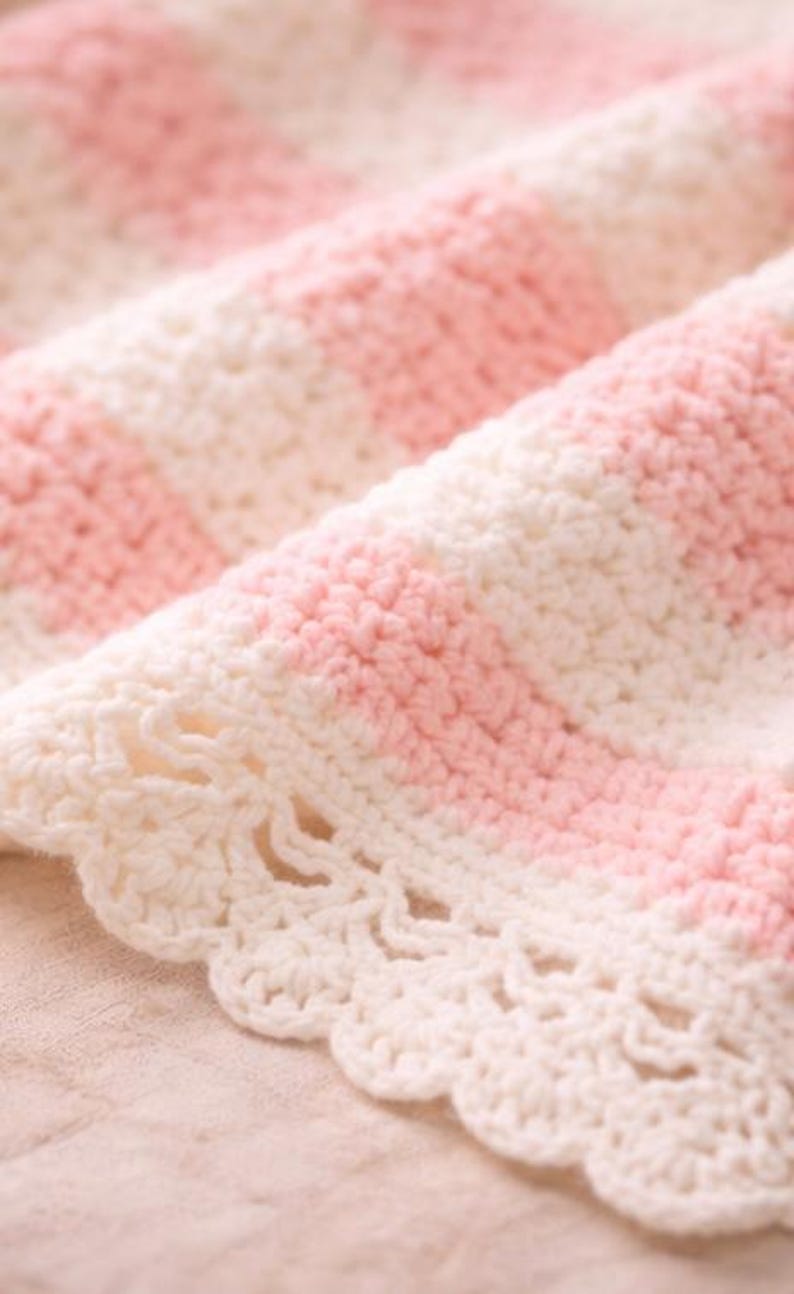 May include: Close-up of a handmade crochet blanket with alternating stripes of soft pink and white. The edge features a delicate, scalloped trim. The blanket is made of a soft yarn, perfect for a baby or as a decorative throw.