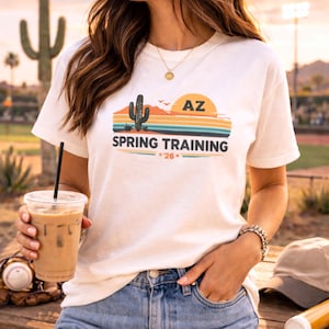 May include: Cream-colored t-shirt featuring a desert landscape design with a cactus, sun, and the text "AZ SPRING TRAINING *26*". The shirt is paired with denim shorts. A baseball, glove, and hat are visible.