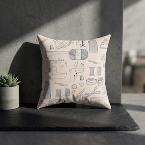 May include: A square beige pillow with a repeating pattern of sewing tools in dark blue. The design includes a sewing machine, spools of thread, scissors, buttons, and a measuring tape.